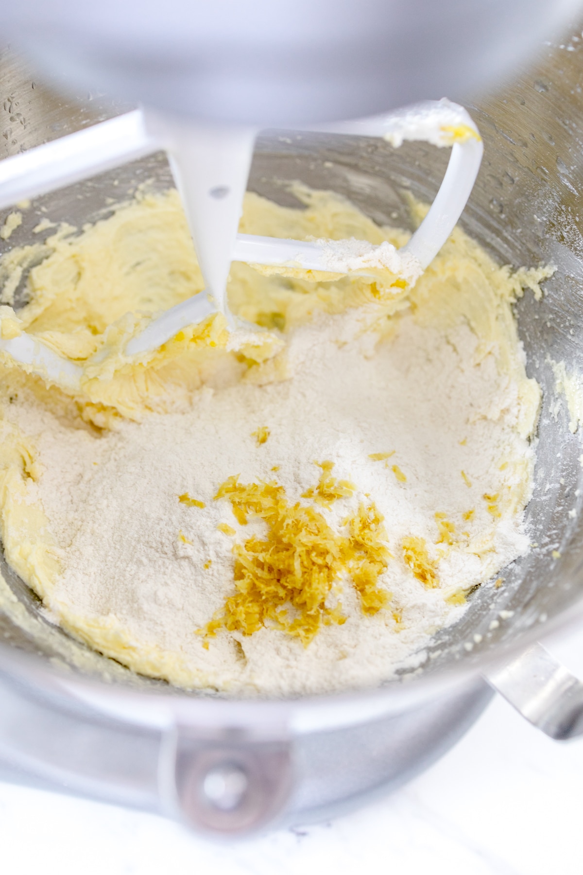 Close up of the bowl of a stand mixer with a creamed mixture in the bottom and a flour mixture with lemon zest on top.