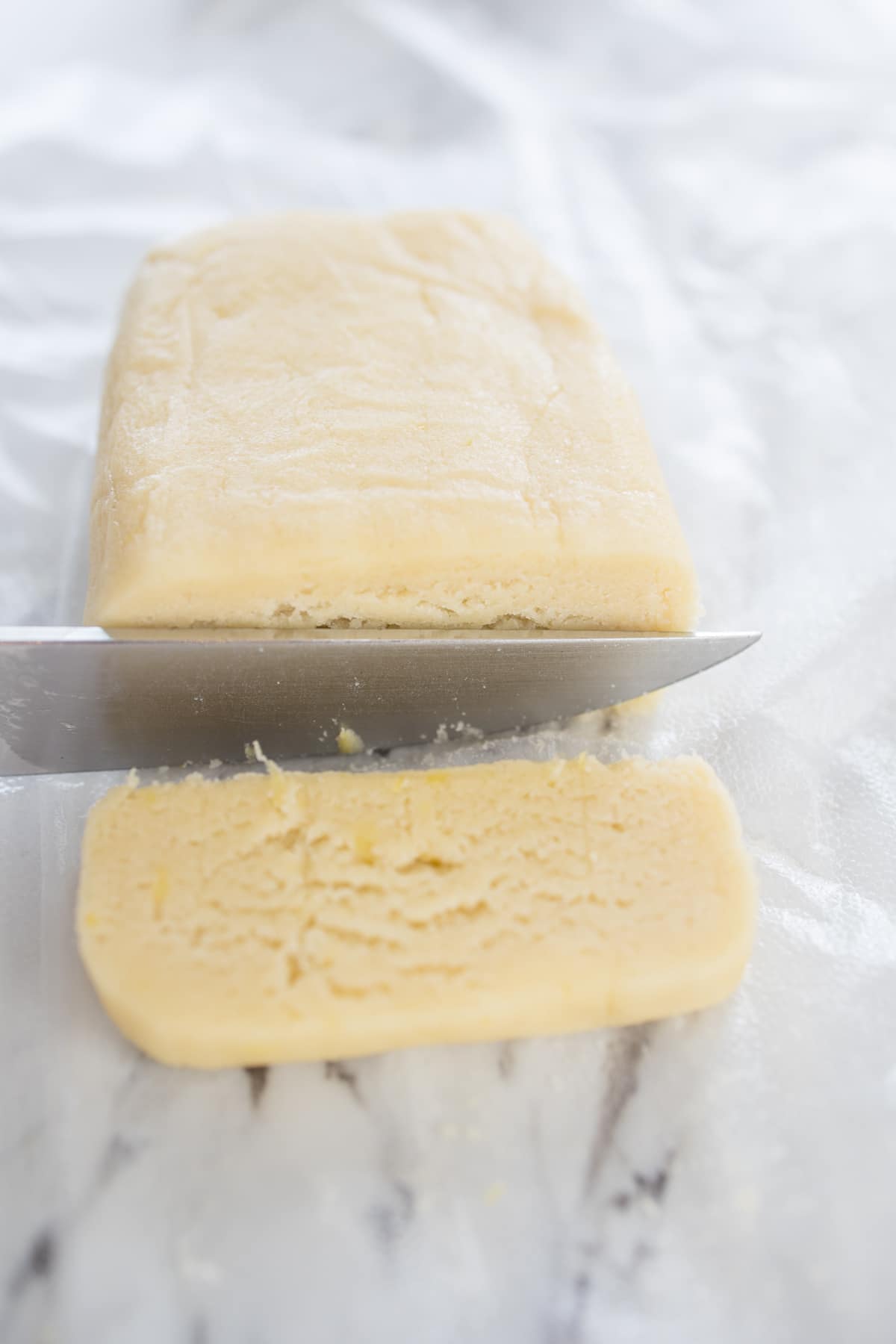 Top view of a slab of lemon shortbread cookie dough being sliced with a sharp knife into thin cookies.