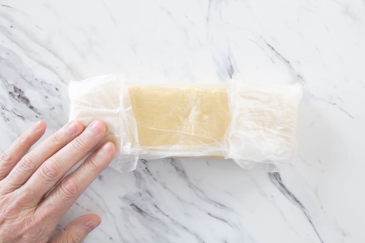 Top view of lemon shortbread cookie dough in a rectangle shape wrapped in plastic wrap with a hand on it.