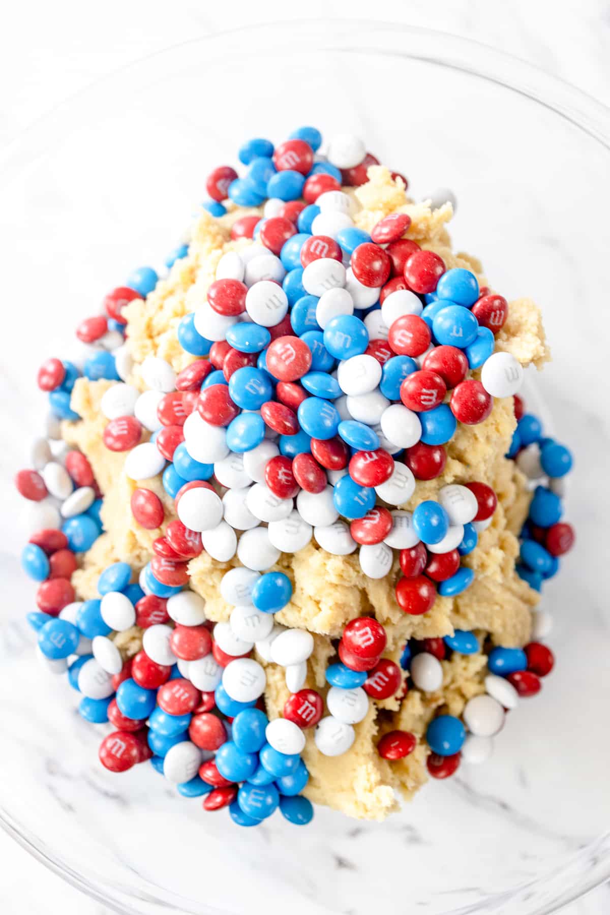 Top view of a glass mixing bowl with cookie dough in the bottom and red, white, and blue M&Ms on top.