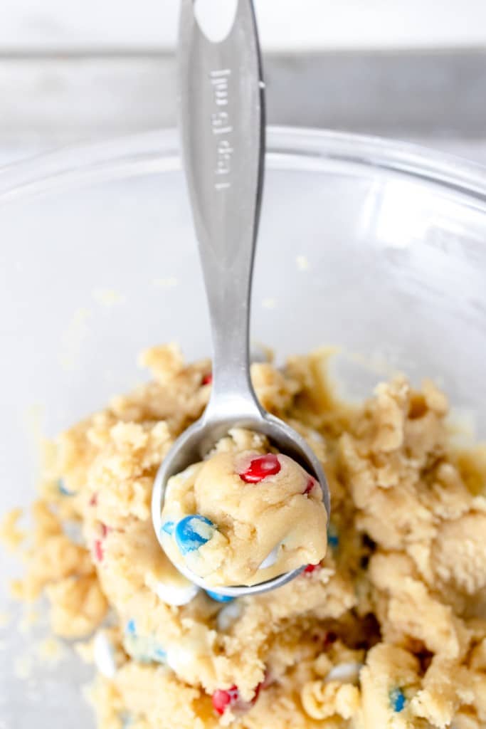 Top view of a glass mixing bowl with Mini M&M cookie doough being scooped by a cookie scoop.