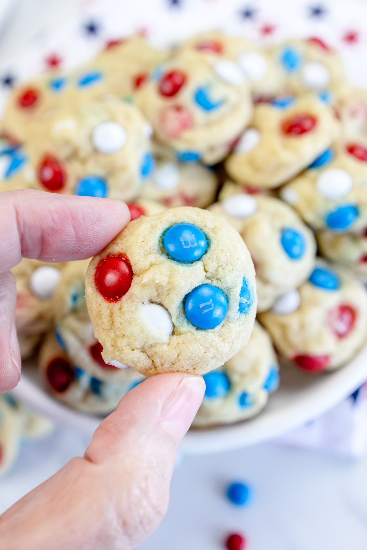 Close up of someone holding a Mini Mini M&M cookie in their hand with more cookies on a plate in the background. 