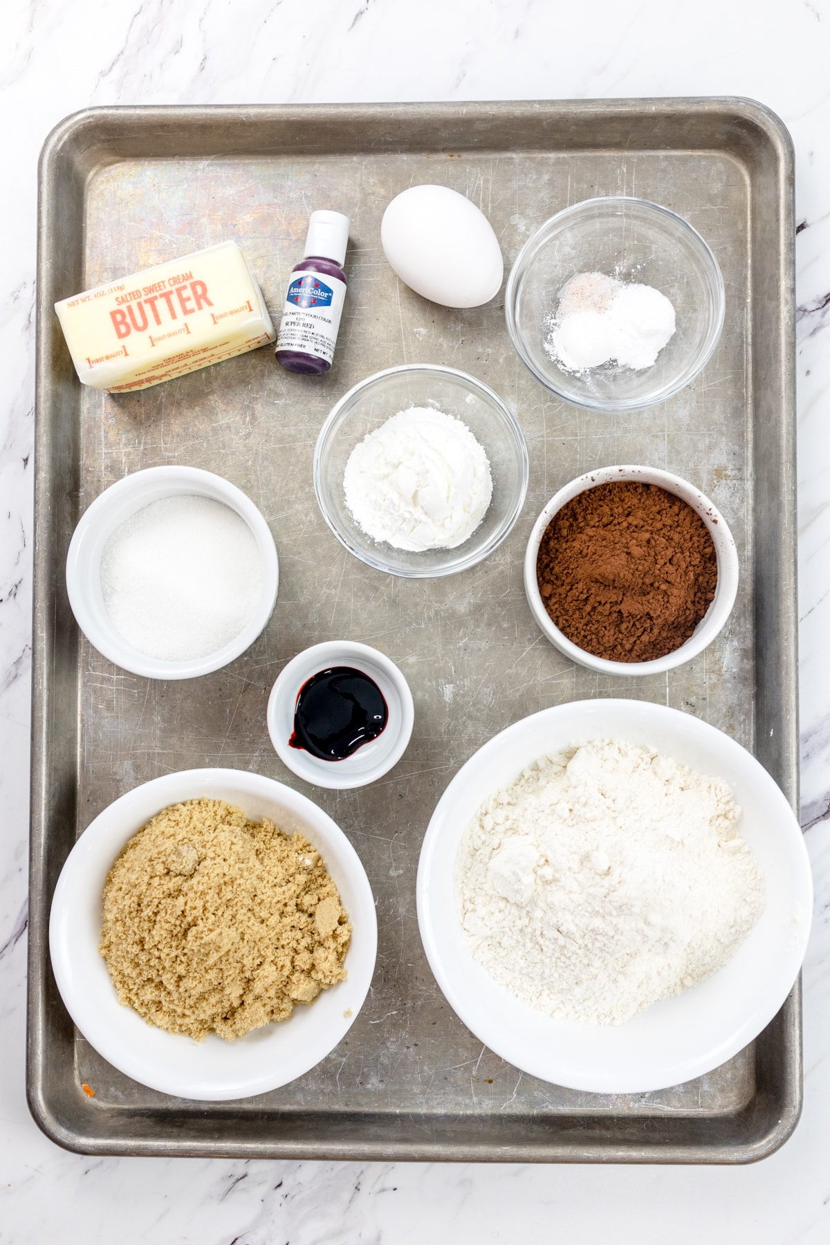 Top view of ingredients needed to make Red Velvet Cookies with Cream Cheese Frosting in small bowls on a baking tray.