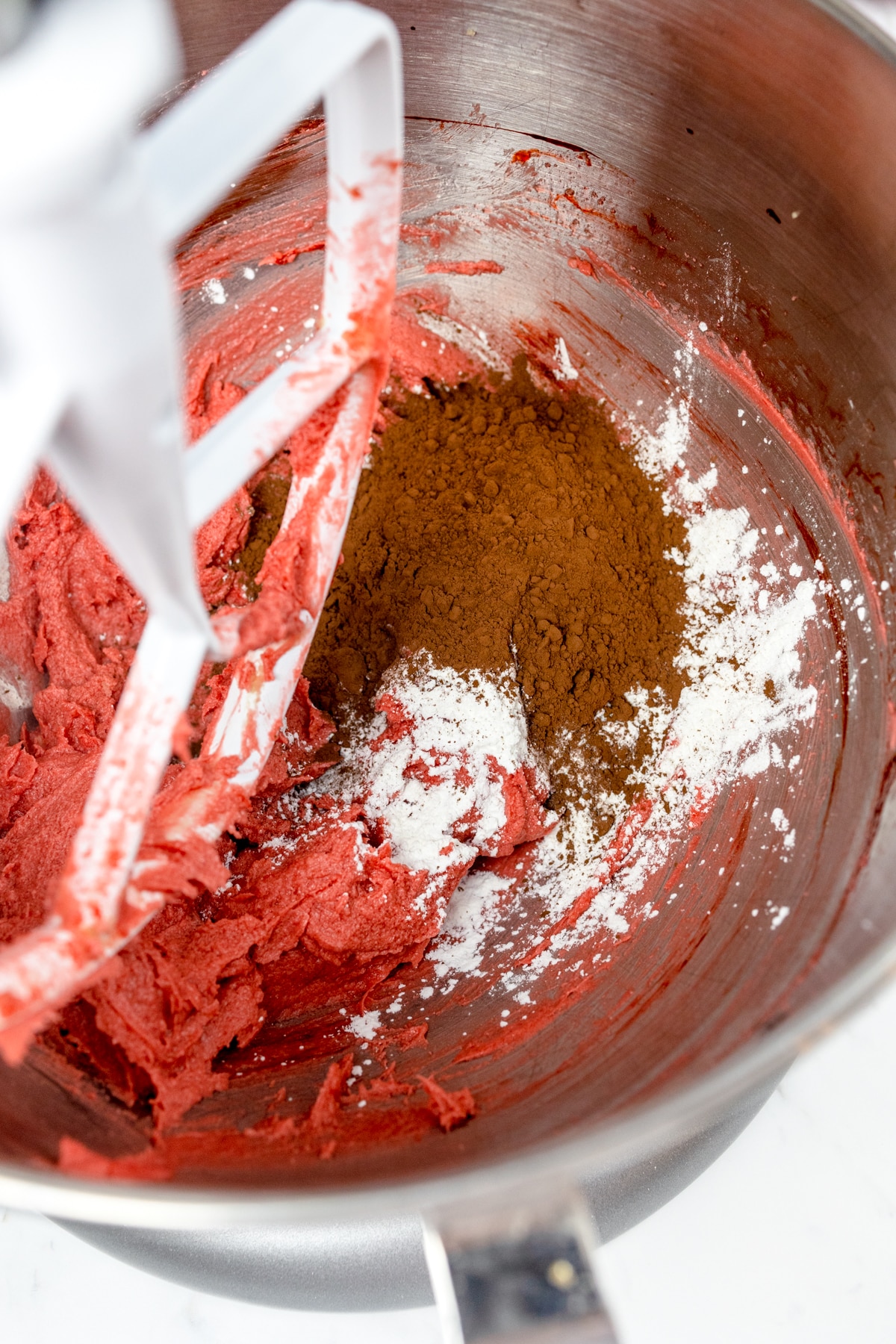 Close up of the bowl of a stand micer with a paddle attachment mixing red food coloring in with cocoa powder and a white powder.