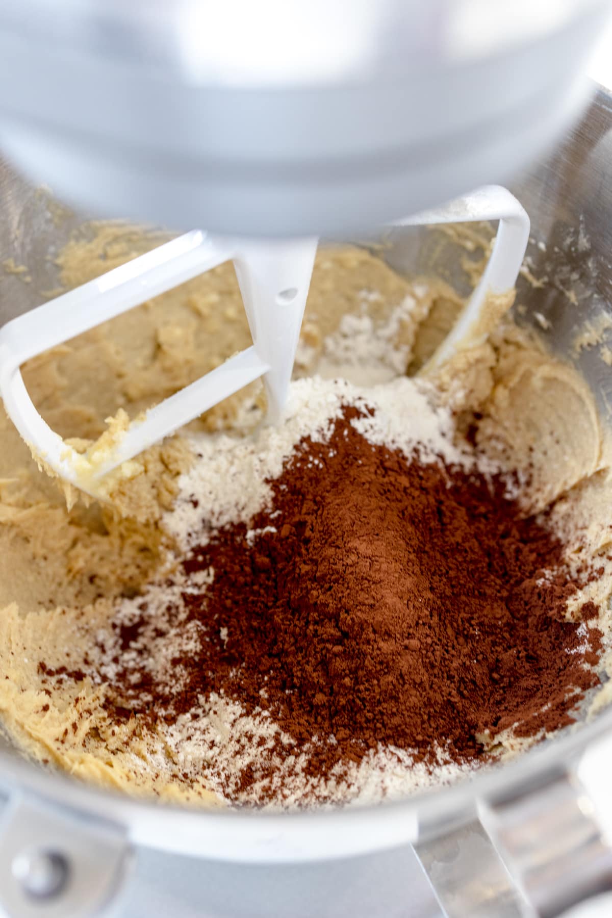 Close up of the bowl of a stand mixer with a paddle attachment mixing cookie dough with cocoa powder on top.