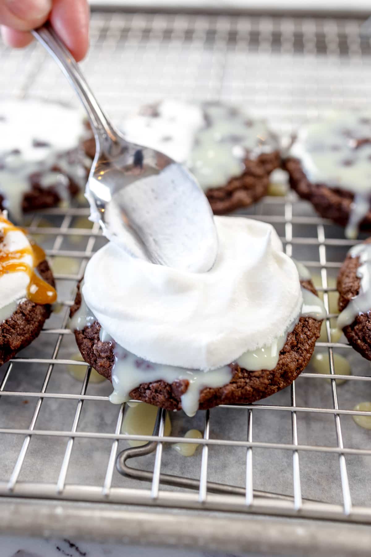 Close up of a spoon patting down whipped cream on top of icing on a chocolate cookie on a wire rack.