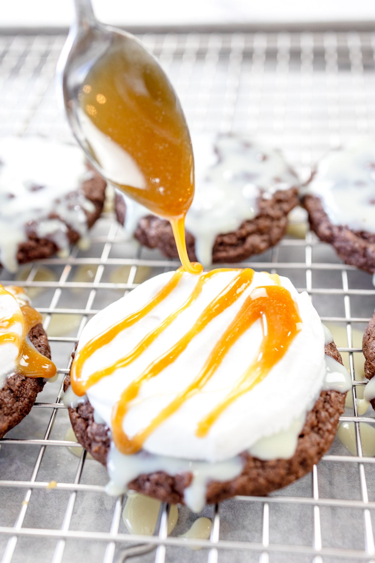 Close up of a spoon drizzling caramel sauce onto icing onto a chocolate cookie on a wire rack.