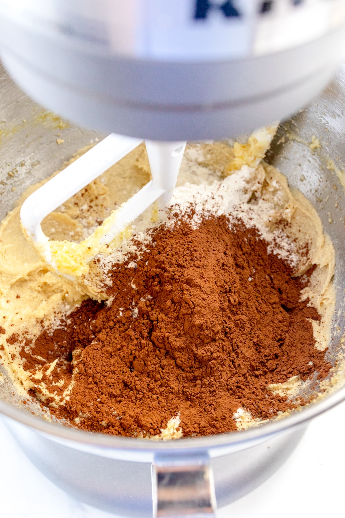 Close up of the bowl of a stand mixer, mixing cookit dough with cooca powder on top with a paddle attachment.