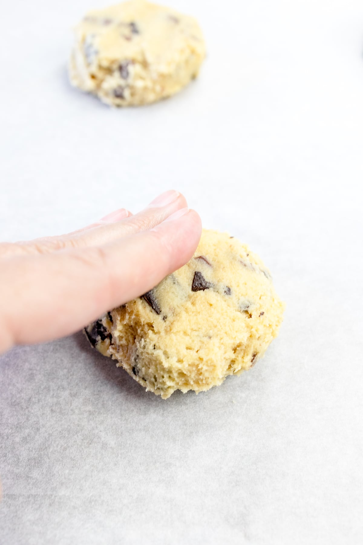 Close up of a ball of cookie dough on parchment pper with fingers pressing it down.