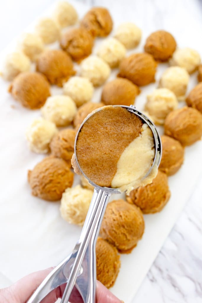 Close up of a cookie scoop scooping both snickerdoodle and gingersnap cookie dough, being held up above balls of cookie dough.