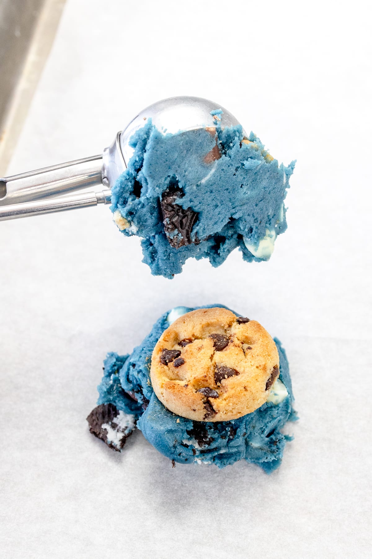 A flattened blue cookie dough ball with a chocolate chip cookie on top sits on a white surface, with anotther blue cookie dough ball being added on top with a cookie scoop.