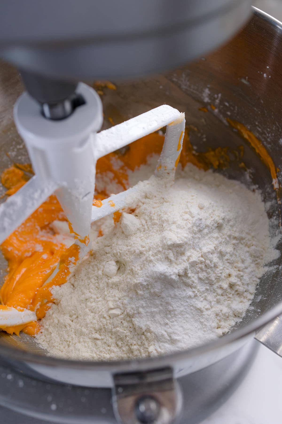 Close up of the bowl of a stand mixer with a paddle attachment in it with an orange mixture and flour mixture being combined.