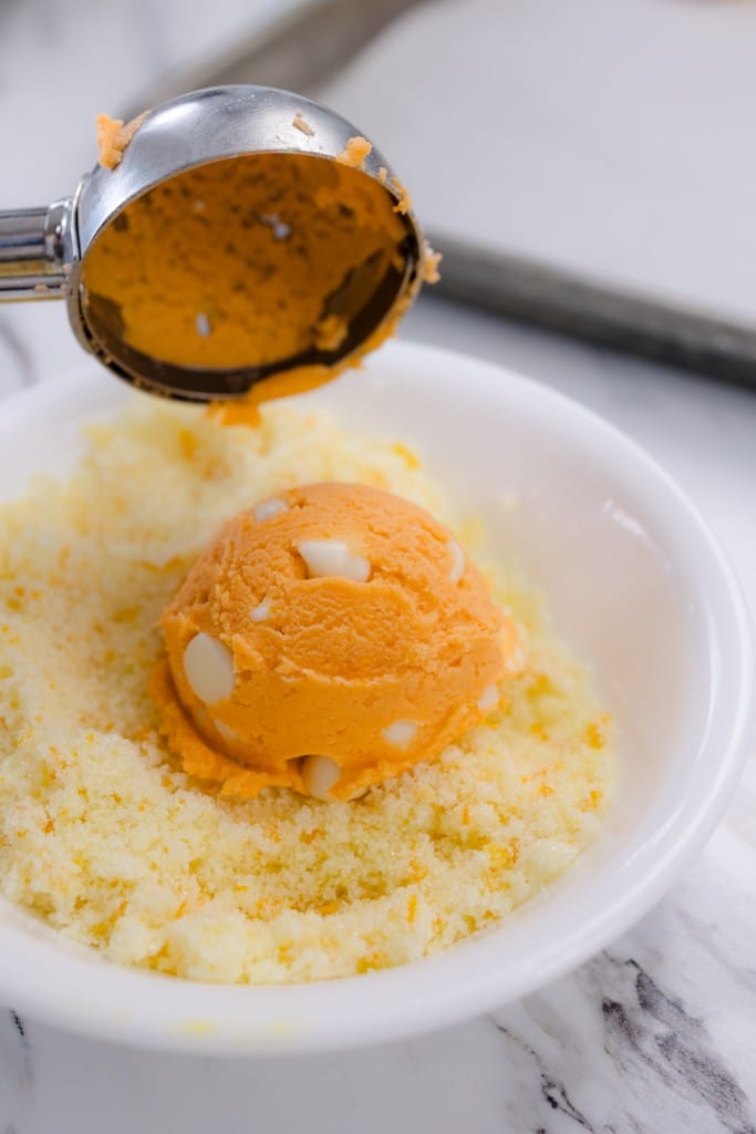 Close up of an orange creamsicle cookie dough ball being placed into a bowl of orange infused sugar by a cookie scoop.