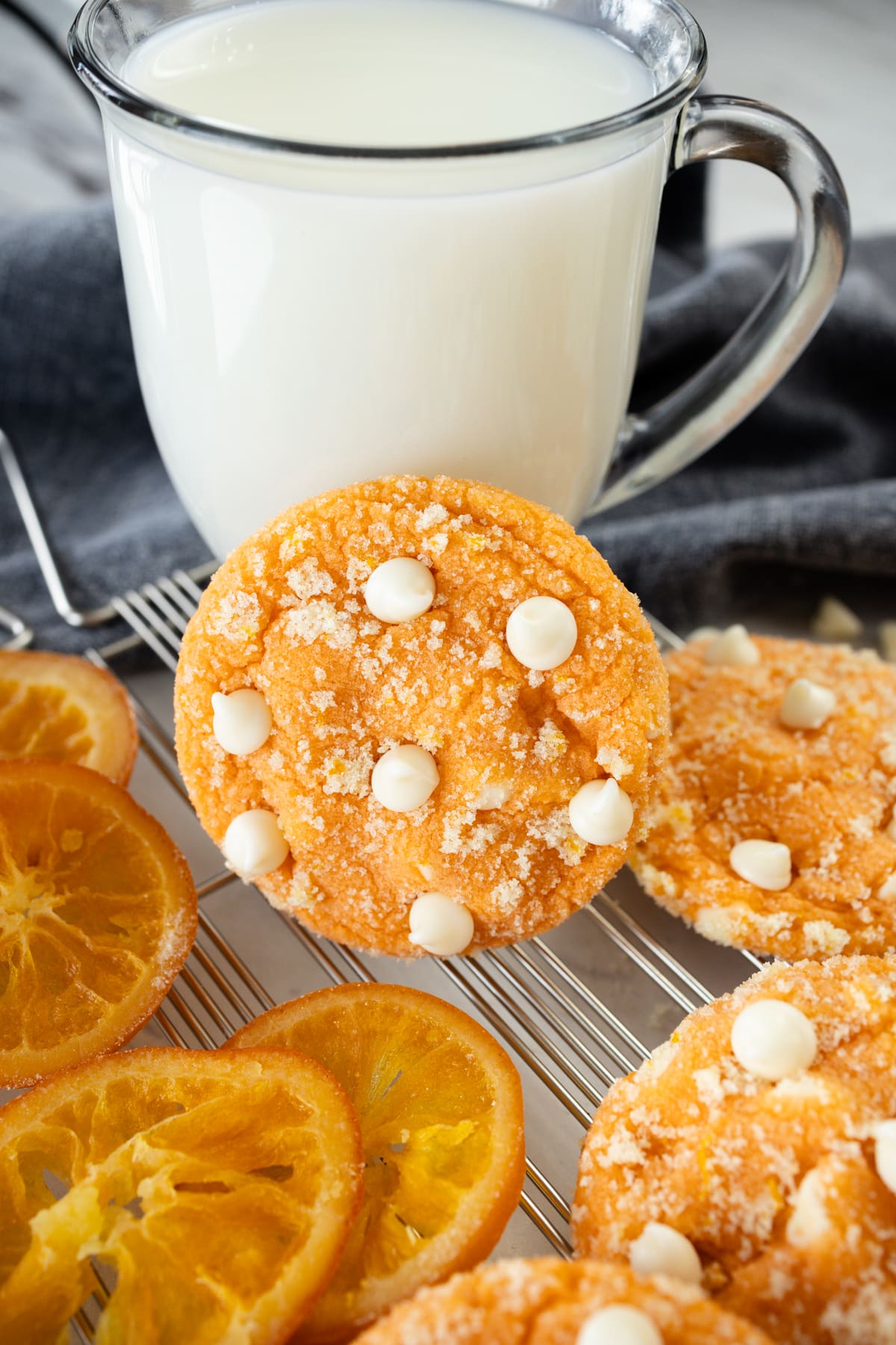 Close up of an orange creamsicle cookie resting on a glass of milk, with orange slics and other cookies surrounding it. 