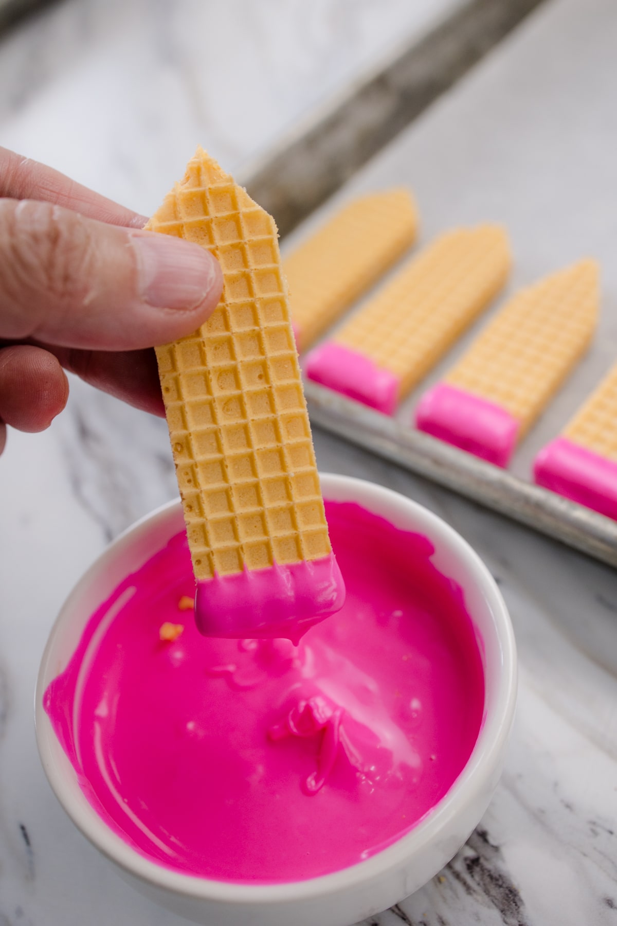 A hand dipping a wafer cookie into melted pink chocolate.