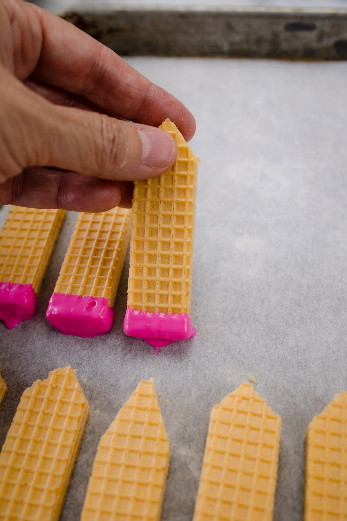 A hand placing a wafer cookie dipped in pink chocolate onto a parchment paper lined baking sheet.