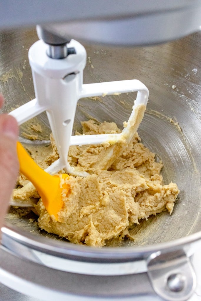 Close up of the bowl of a stand mixer with a paddle attachment and cookie dough in it, with a rubber spatula scraping down the sides of the bowl.