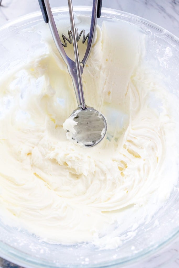 Top view of a glass mixing bowl with white frosting in it and a cookie scoop scooping out some frosting.