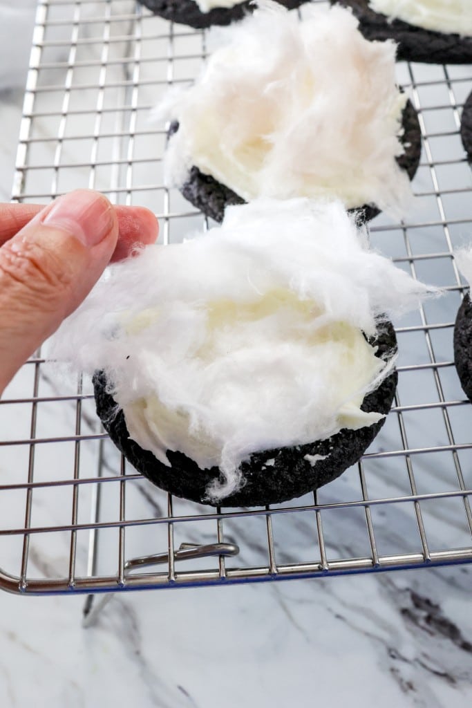 Close up of chocolate cookies with frosting on a wire rack being topped with white candy floss.