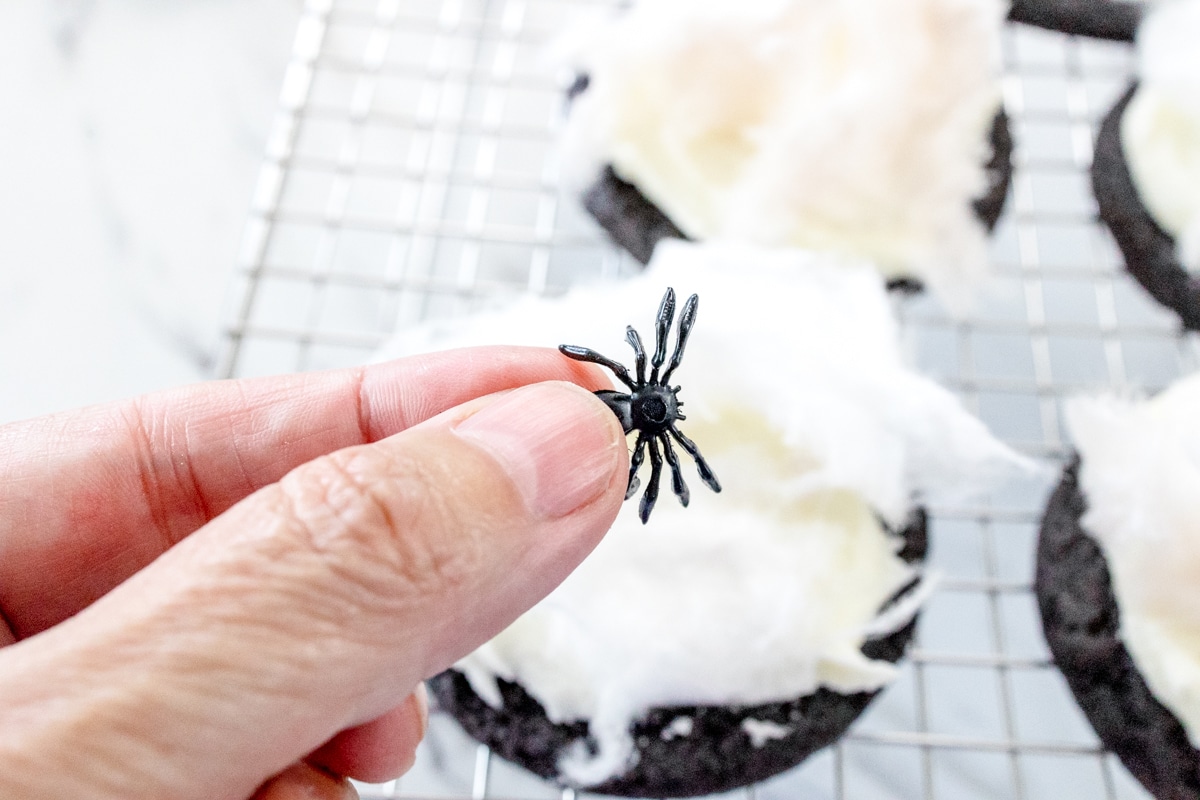 Top view of frosted Halloween Spider Web Cookies on a wire rack with someone holding a plastic spider above it.