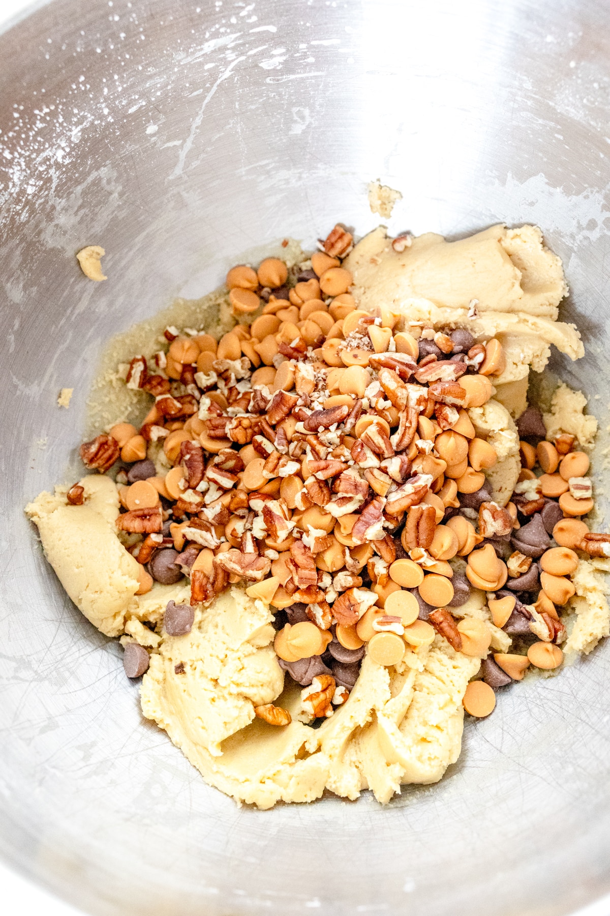 Close up of a bowl of pecans, chocolate chips, and butterscotch chips on top of the cookie dough, ready to be mixed together.