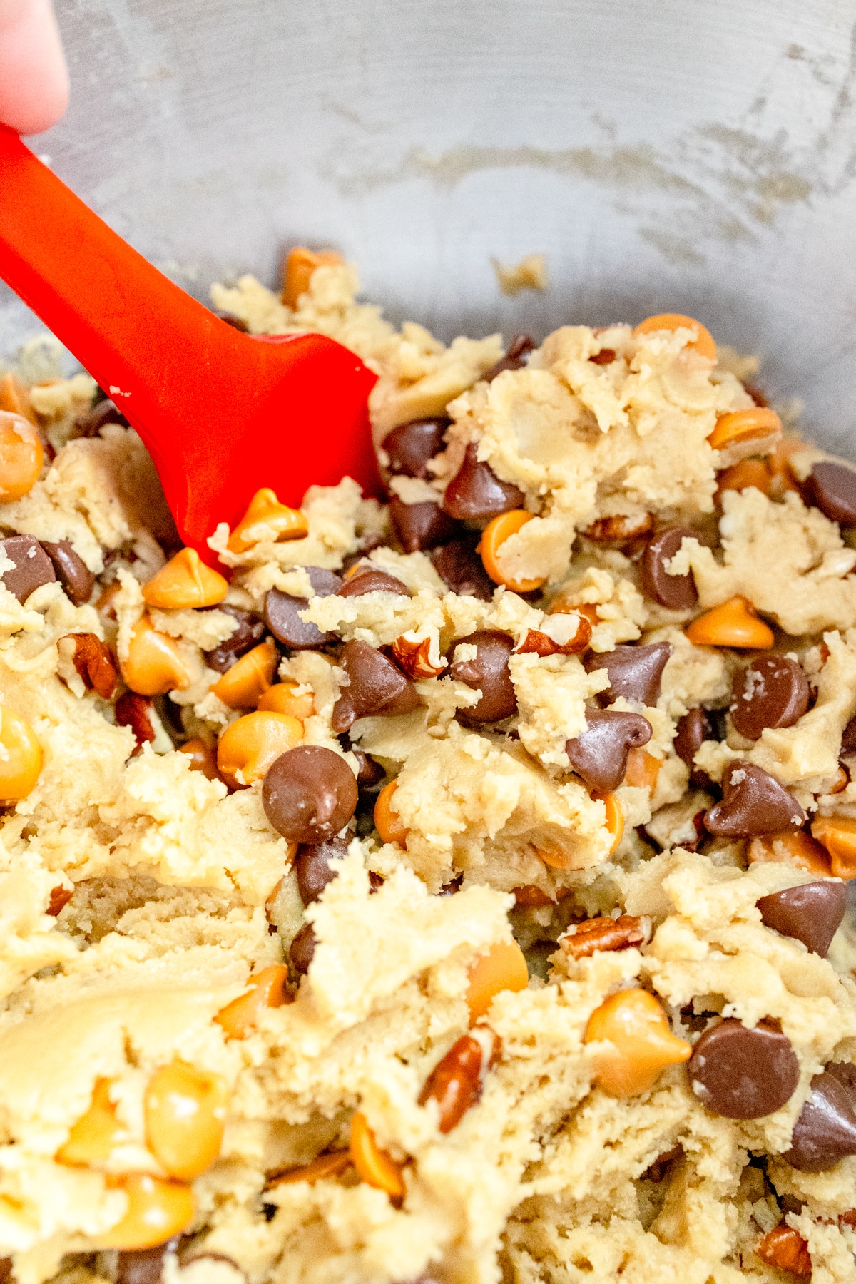 Close up of a bowl of pecans, chocolate chips, and butterscotch chips on top of the cookie dough, being mixed together by hand with a spatula.