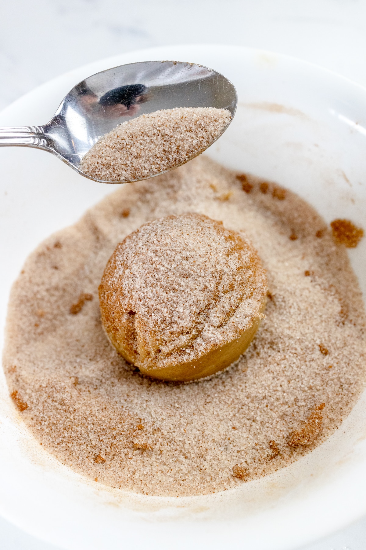 Close up of a cookie dough ball being rolled into a small bowl with cinnamon sugar in it.