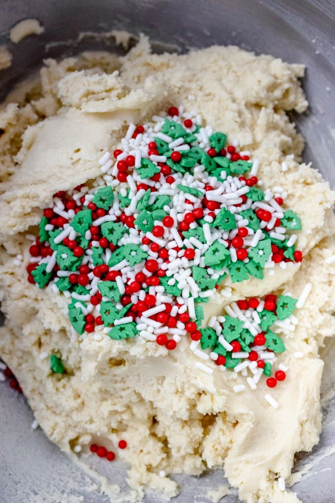Close up of the bowl of a stand mixer that shows red, green and white sprinkles on top of cookie dough, ready to be mixed together.