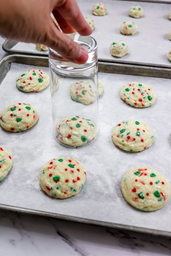 Top view of freshly baked Christmas Sugar Cookies on a baking tray lined with parchment paper, and a glass tumbler is being placed around one to make it into a perfect circle.