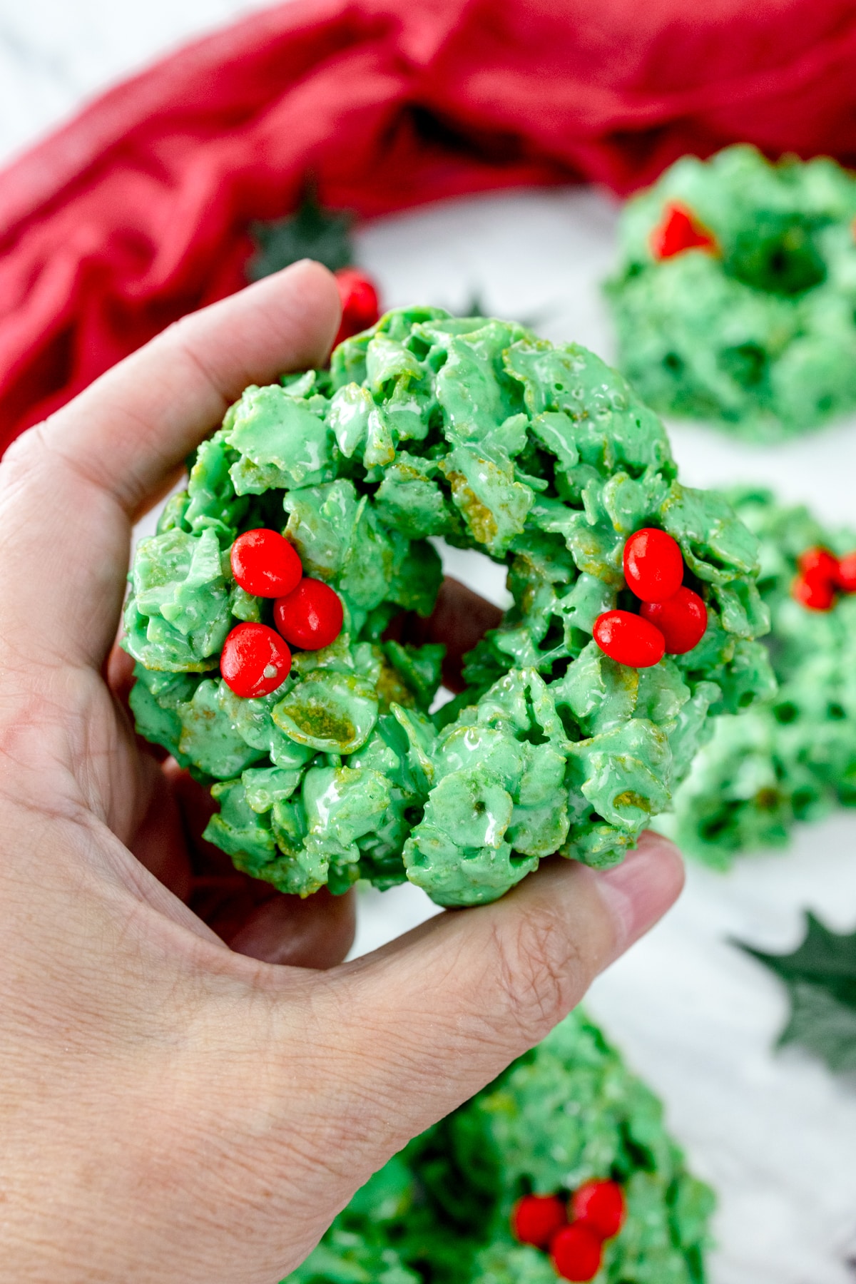 Close up view of a hand holding a Christmas Wreath Cookie above a white surface with other Christmas Wreath Cookies and Christmas decorations.