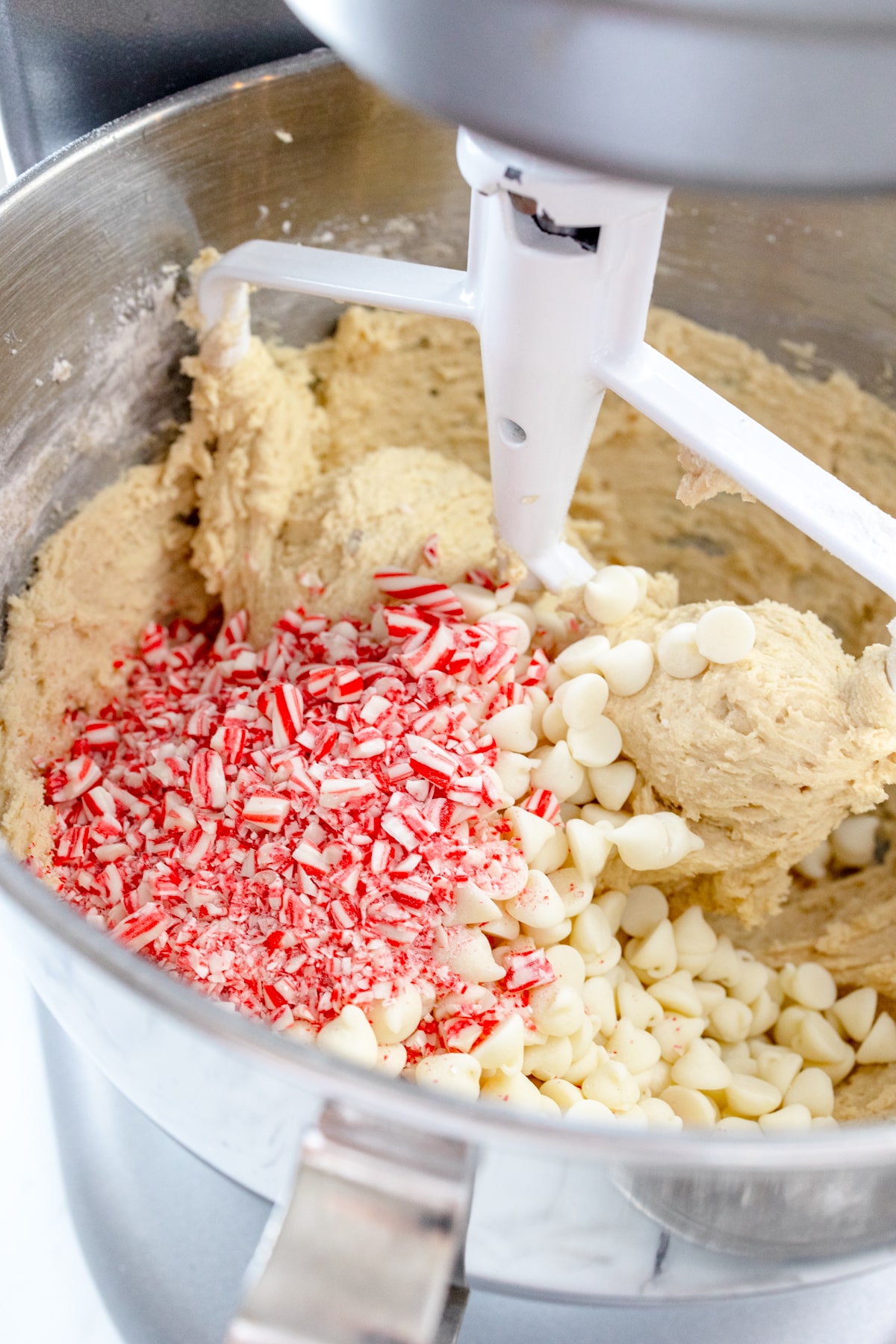 Close up of the bowl of a stand mixer with cookie dough in it with chocolate chips and crushed candy canes that have been added on top to mix in.