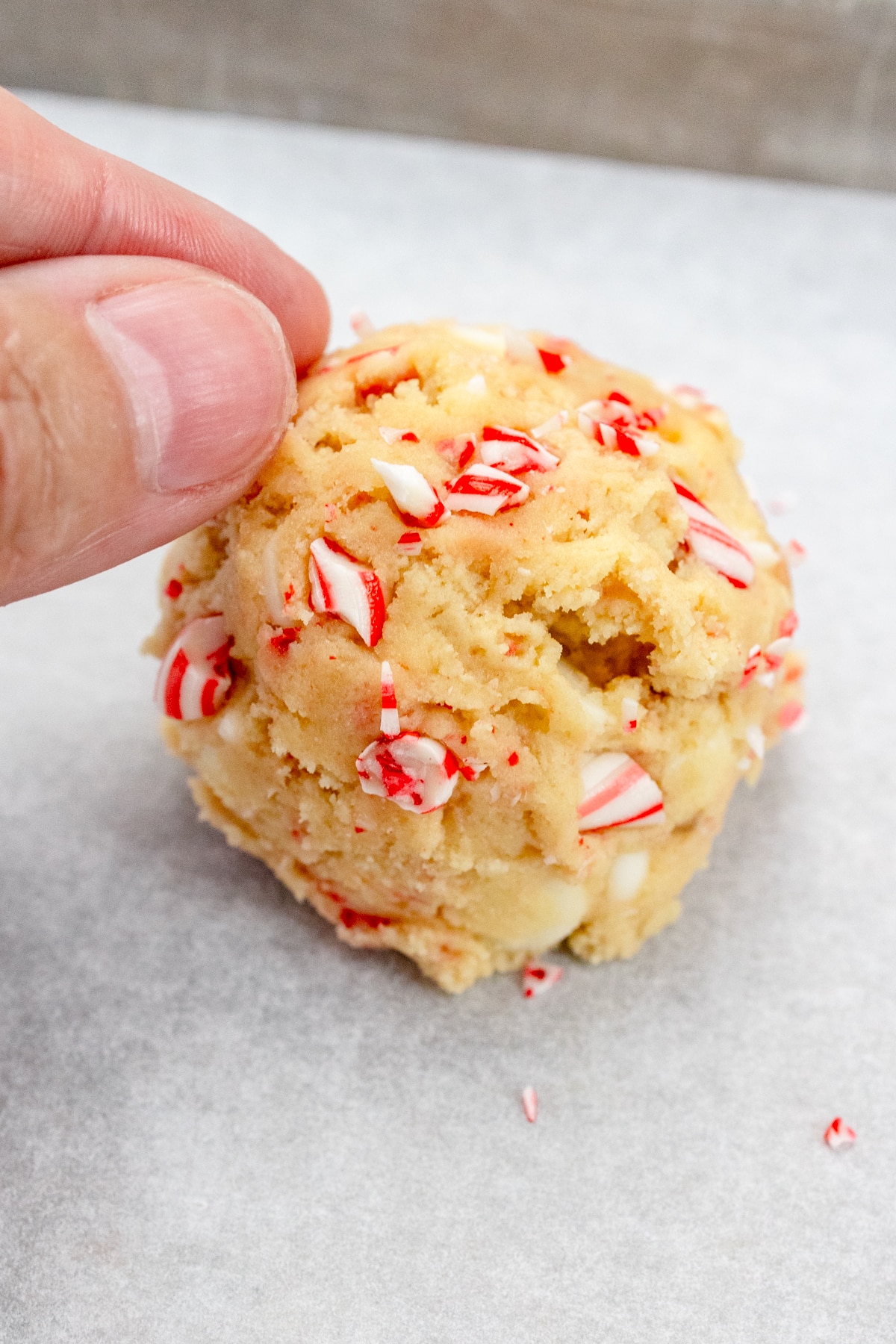 Close up view of fingers adding more crushed candy pieces to the top of a Close up of a scoop of cookie dough on a baking tray lined with parchment paper.