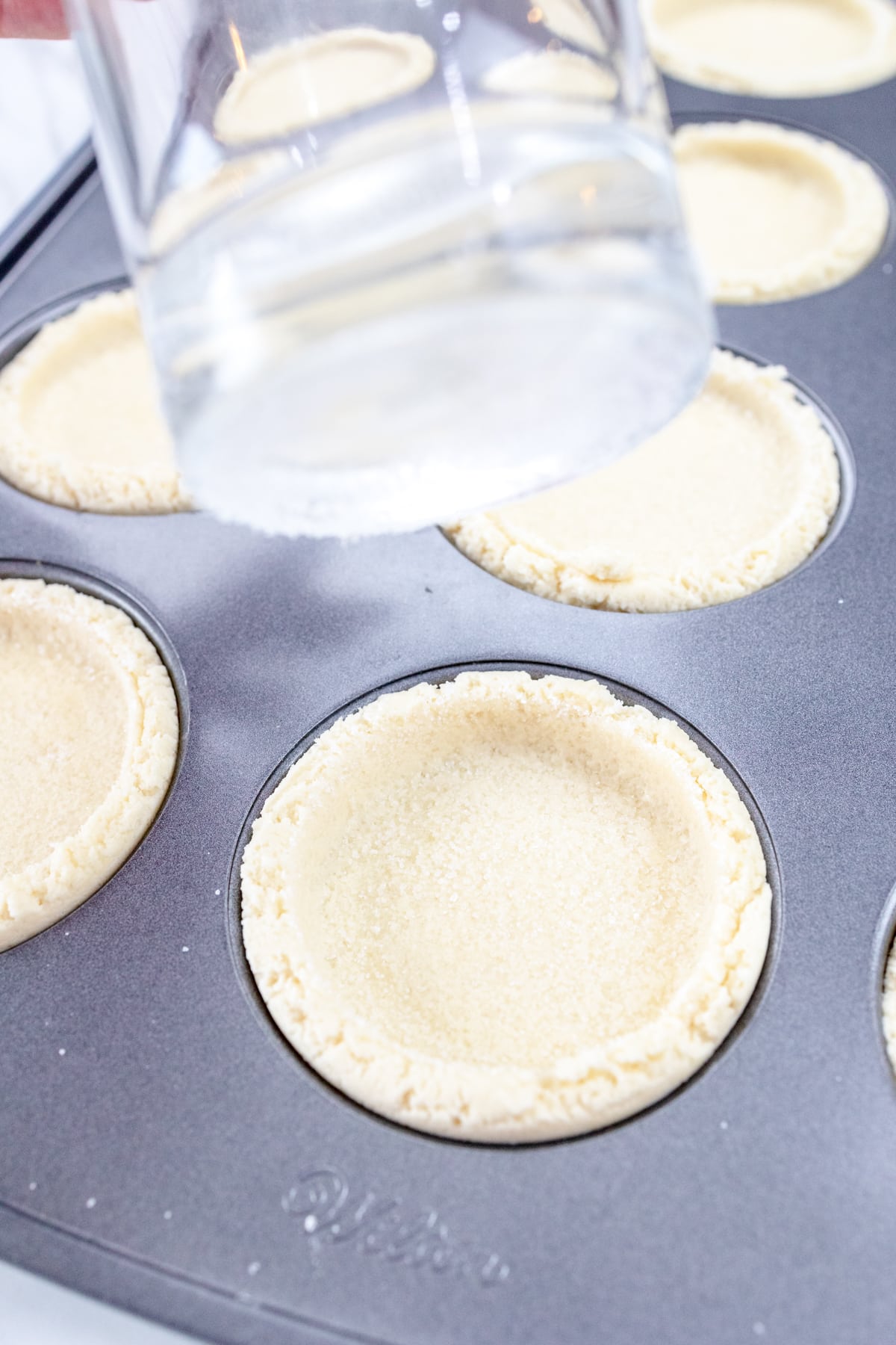 Close up of the bottom of a glass that has been dipped in sugar, being lifted from having just pressed the cookie dough down flat into the muffin top pan.