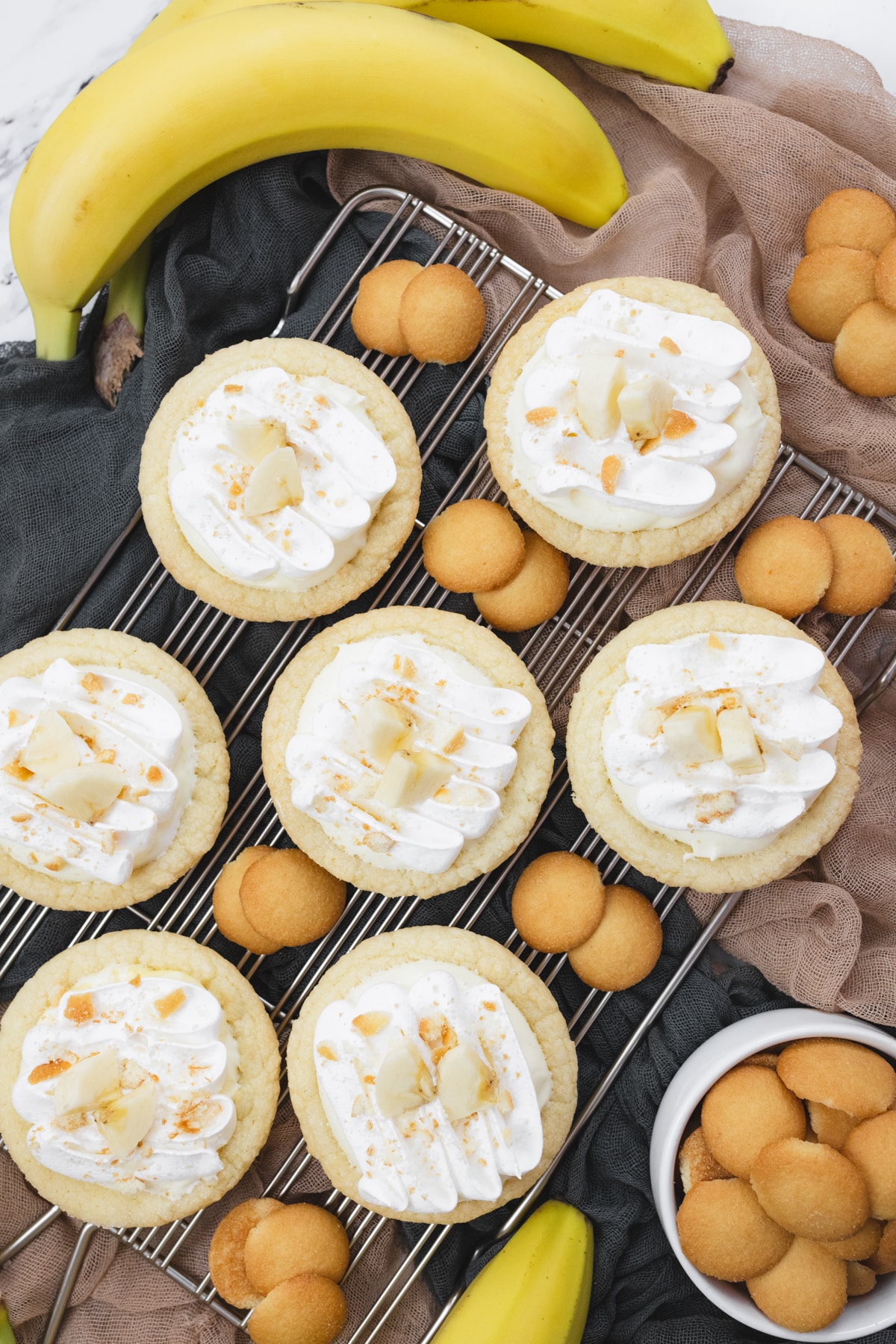 Top view of finished Banana Cream Pie Cookies on a wire rack garnished with banana slices and sprinkled with mini nilla wafer crumbles, surrounded by mini nilla wafers and bananas.