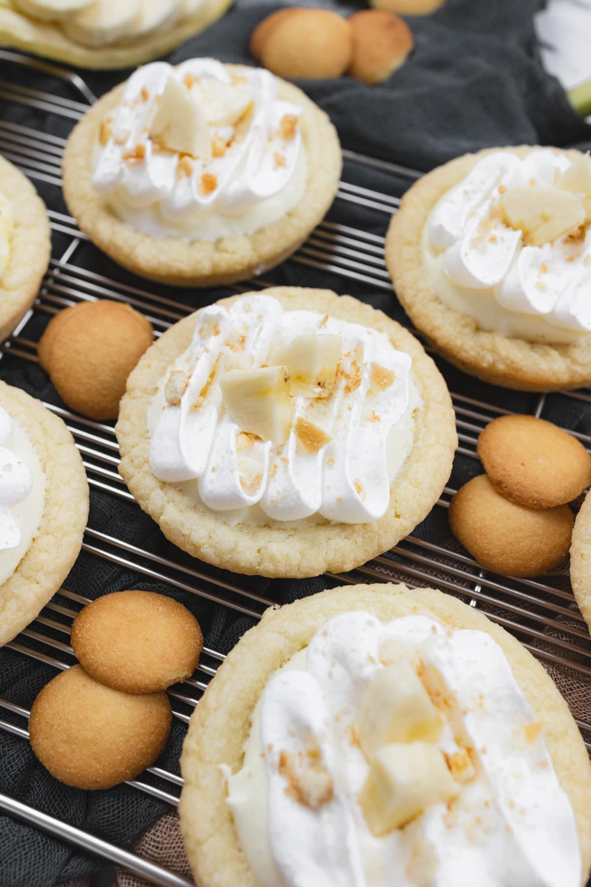 Close up of finished Banana Cream Pie Cookies on a wire rack garnished with banana slices and sprinkled with mini nilla wafer crumbles, surrounded by mini nilla wafers and bananas.