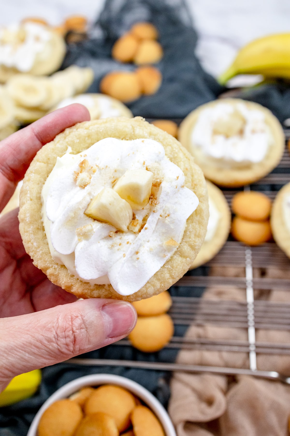 Close up of a hand holding a finished Banana Cream Pie Cookies garnished with banana slices and sprinkled with mini nilla wafer crumbles, in front of a table with other cookies on it surrounded by mini nilla wafers and bananas.