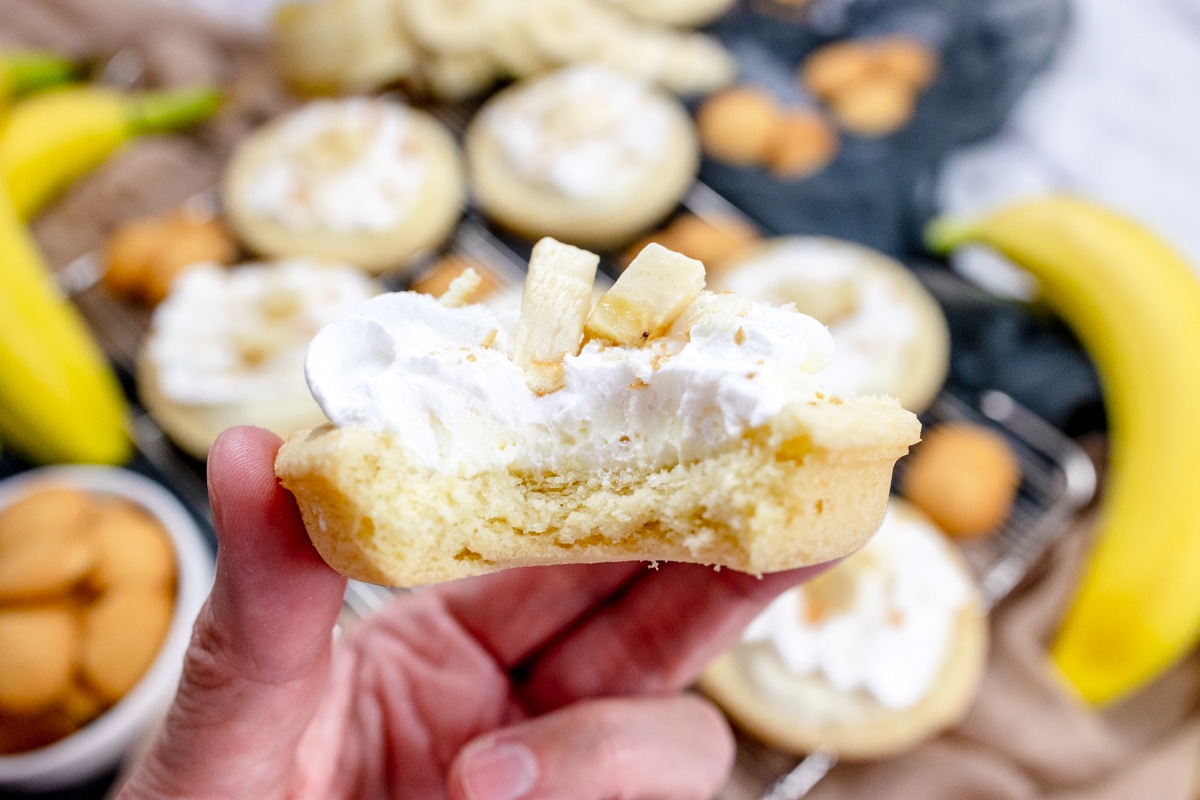 Close up of a hand holding a finished Banana Cream Pie Cookies garnished with banana slices and sprinkled with mini nilla wafer crumbles, in front of a table with other cookies on it surrounded by mini nilla wafers and bananas.