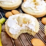 Close up of finished Banana Cream Pie Cookies on a wire rack garnished with banana slices and sprinkled with mini nilla wafer crumbles, surrounded by mini nilla wafers and bananas. The foremost cookie has a bite taken from it.