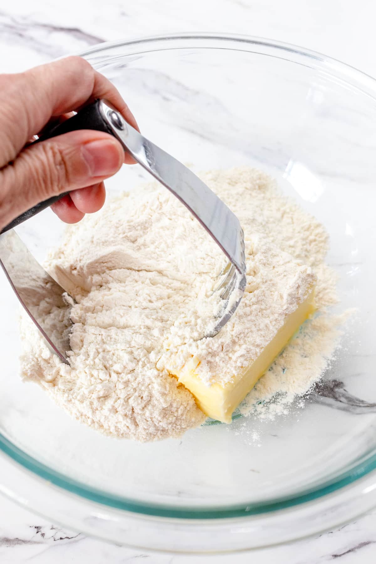 Close up of a hand blending butter and all-purpose flour in a mixing bowl with a pastry blender.