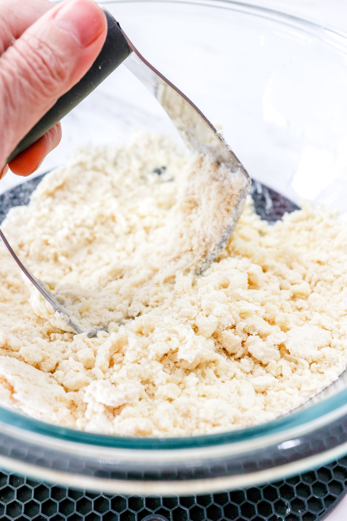 Close up of a hand blending butter and all-purpose flour in a mixing bowl with a pastry blender.