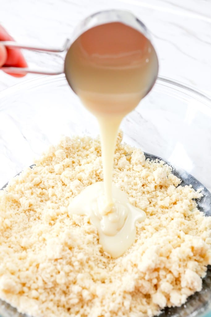 Close up of a hand holding a measuring cup which is pouring condensed milk over blended butter and all-purpose flour in a mixing bowl.