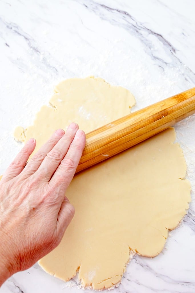 Close up of a hand rolling out cookie dough with a rolling pin on a counter sprinkled with a light dusting of flour.