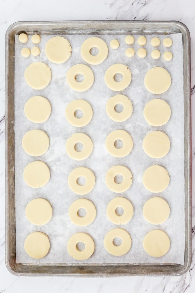 Top view of a baking tray lined with parchment paper with unbaked cookies on it, half are full-circles and half have holes cut out from the center of them, and the dough from the holes is also on the tray.