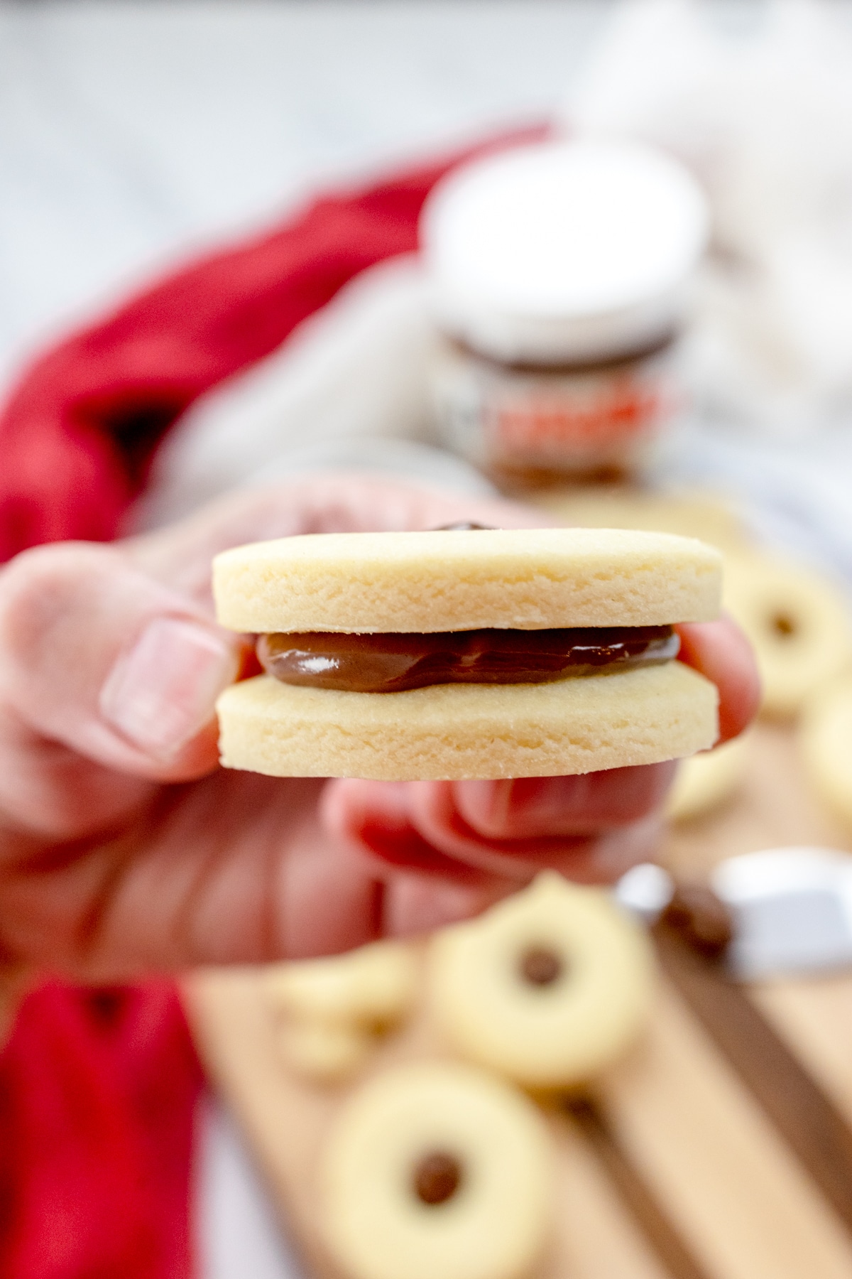 Close up and side-view of a hand holding a finished cookie with a dollop of chocolate sandwiched between two cookies.