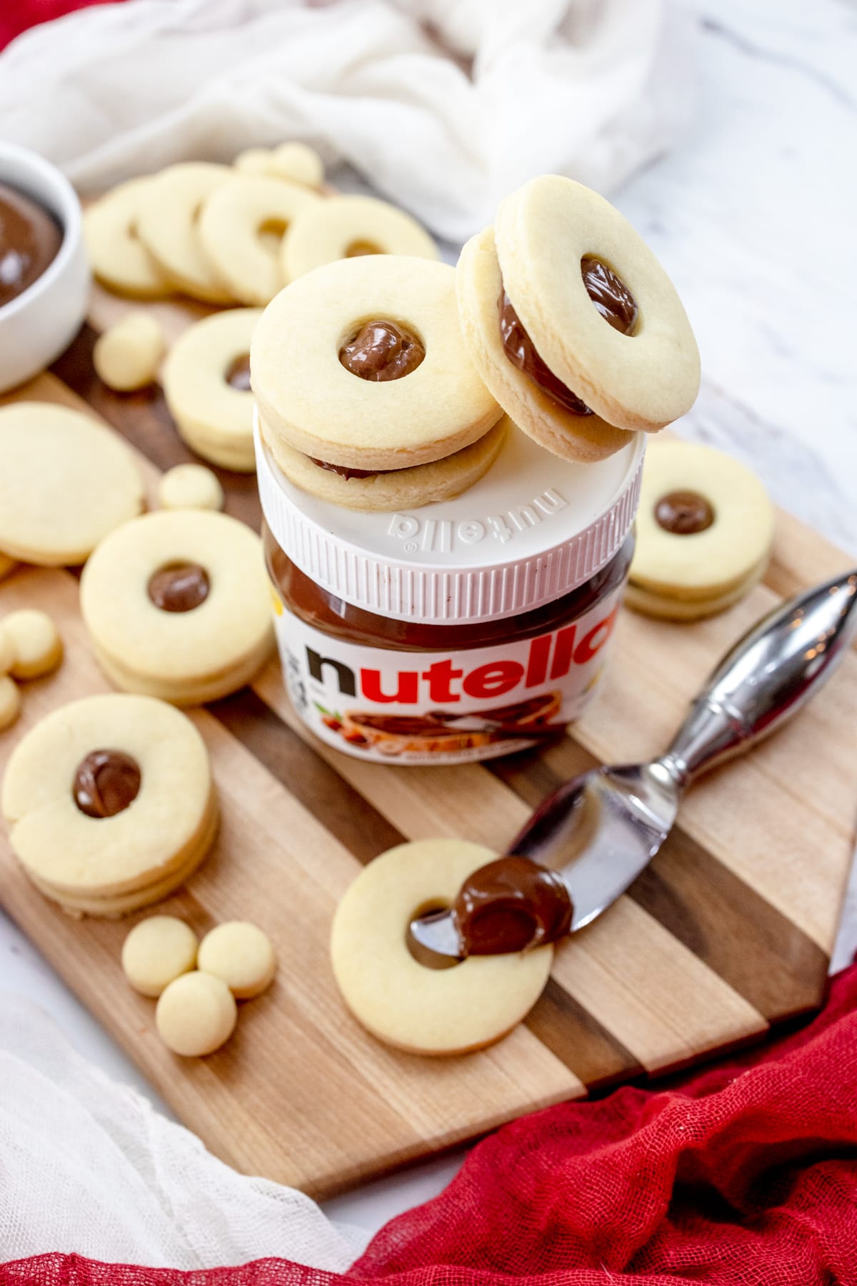 Close up of a prepping area that has a chopping board, Nutella jar with two finished cookies on top, a bowl of nutella, a knife with chocolate spread on it, with unfinished and finished baked cookies around them.