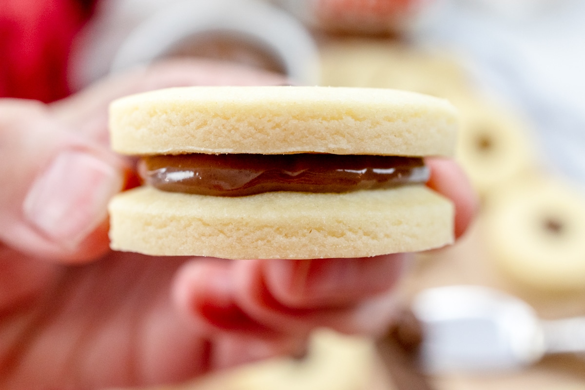Close up and side-view of a hand holding a finished cookie with a dollop of chocolate sandwiched between two cookies.