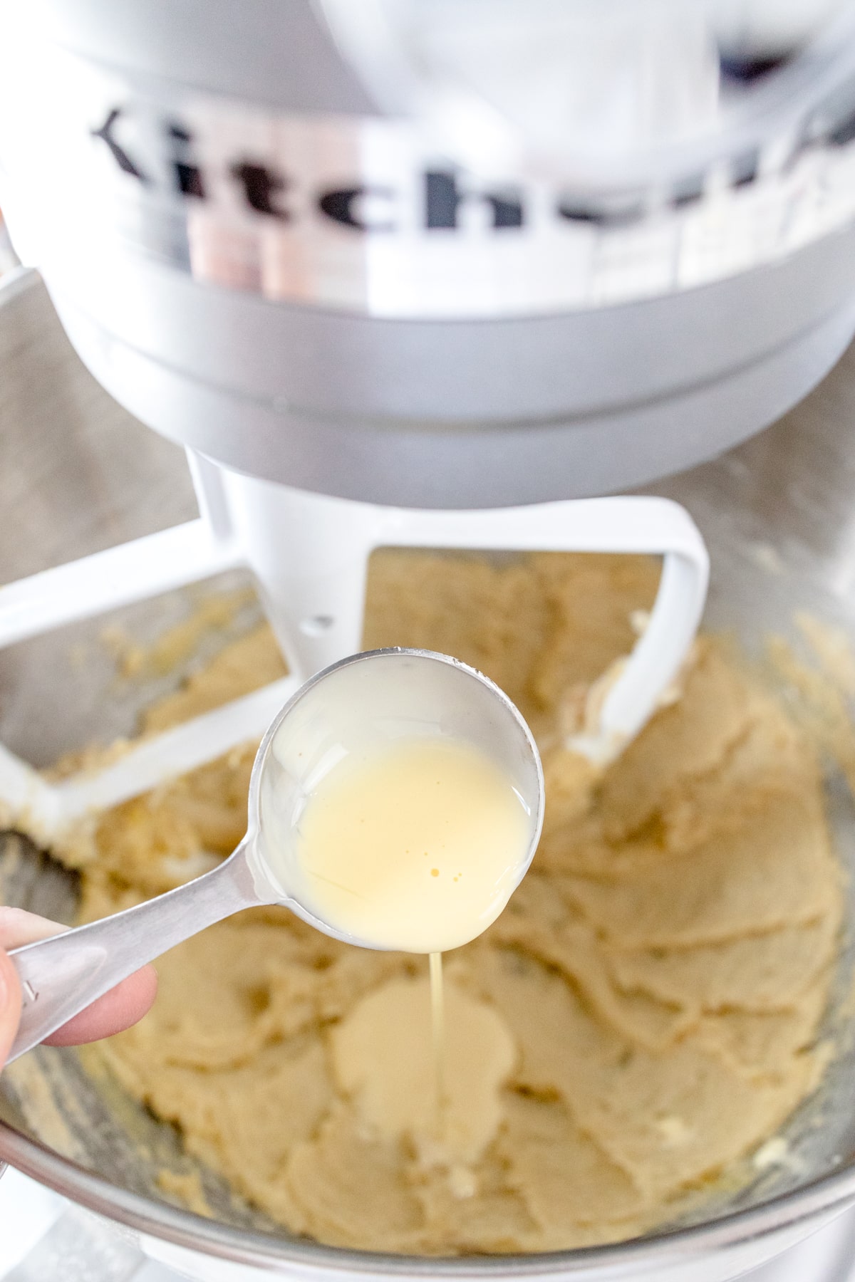 Close up of the bowl of a stand mixer with a creamed mixture in it with eggnog being poured on top.