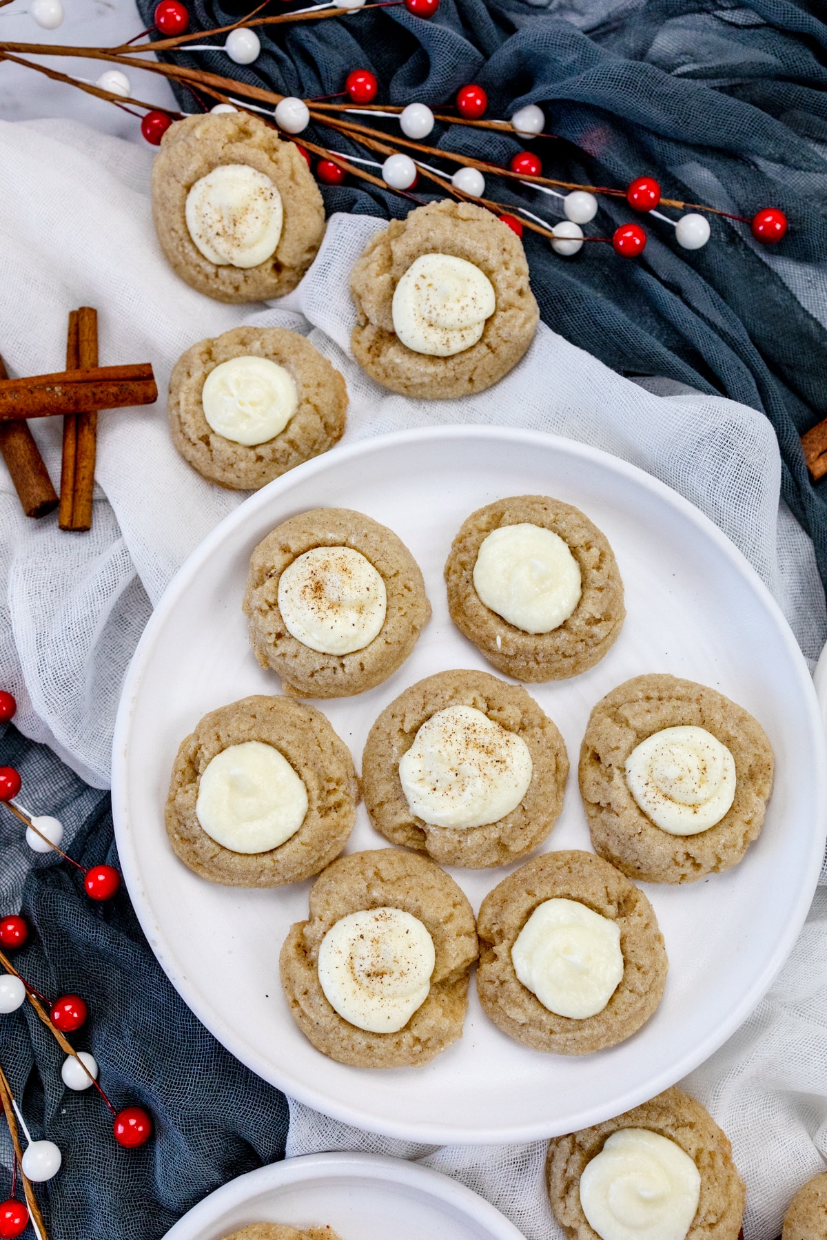 Top view of Eggnog Thumbprint Cookies on a white plate with other cookies and Christmas decorations around it.