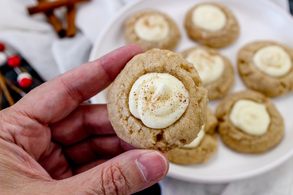 Close up of a hand holding an Eggnog Thumbprint Cookie above a plate of Eggnog Thumbprint Cookies.