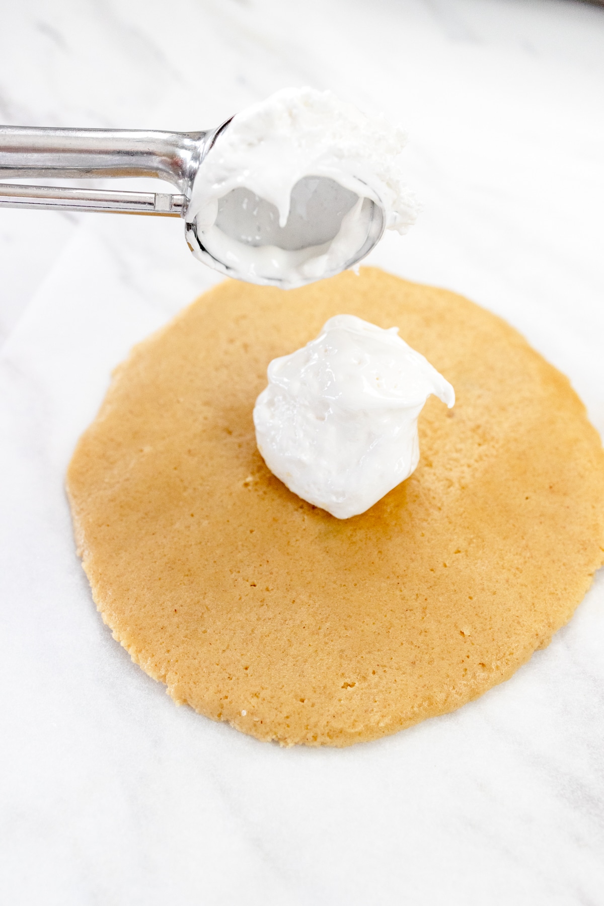 Close up of a cookie scoop scooping marshmallow fluff onto a flattened cookie dough ball.